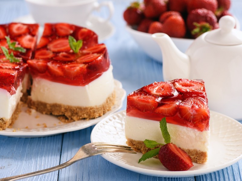 A piece of no-bake strawberry cheesecake on a plate. In the background, the rest of the cake, a teapot and a bowl of strawberries can be seen blurred.