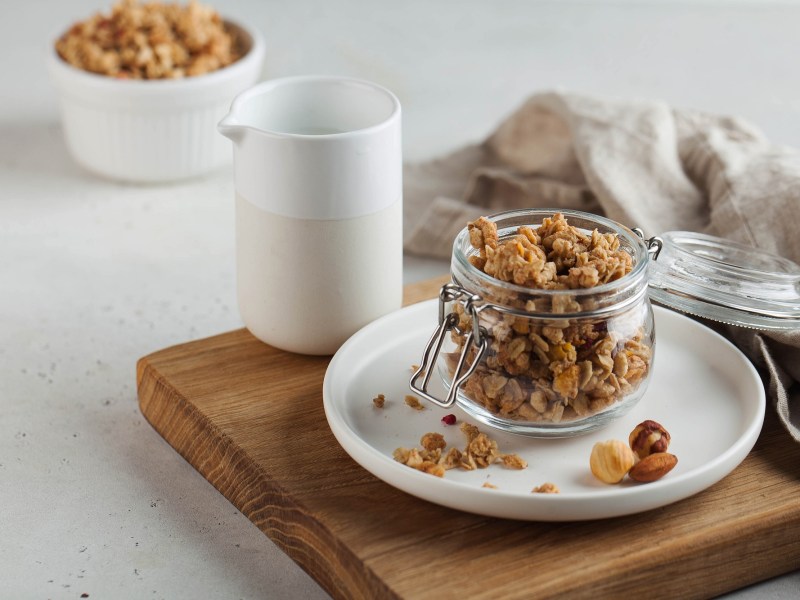 A clear glass sits on a table. Inside the glass, there is crunchy granola. The granola is golden brown and made of oats, nuts, and seeds. Some pieces are small, others are bigger. You can see the different textures.