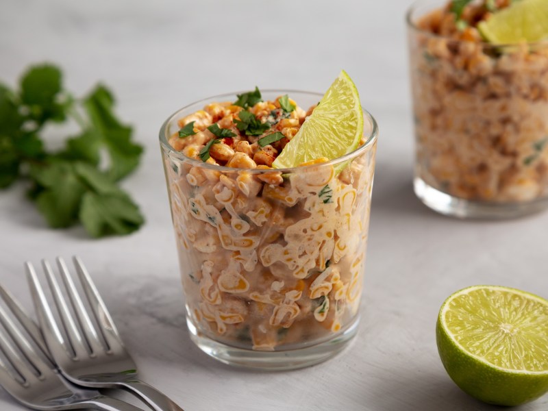 2 glasses of esquites, Mexican street corn salad, garnished with lime wedges and cilantro, next to two forks, fresh cilantro, and a halved lime.