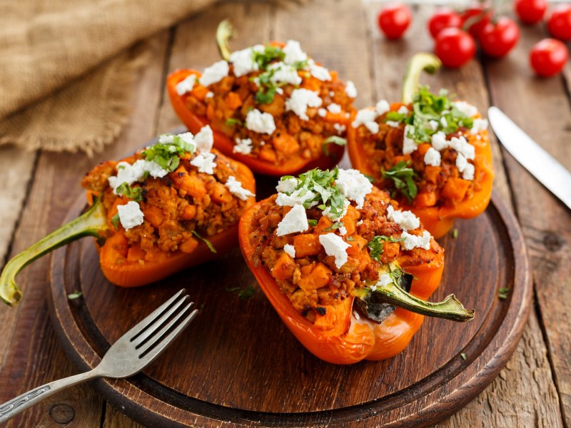 Four halves of grilled stuffed bell peppers lie on a wooden board. Cutlery lies next to it.