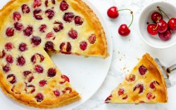 An overhead view of a Pudding-Cherry Cake with a slice cut out. The slice of cake and a bowl of fresh cherries are next to it.