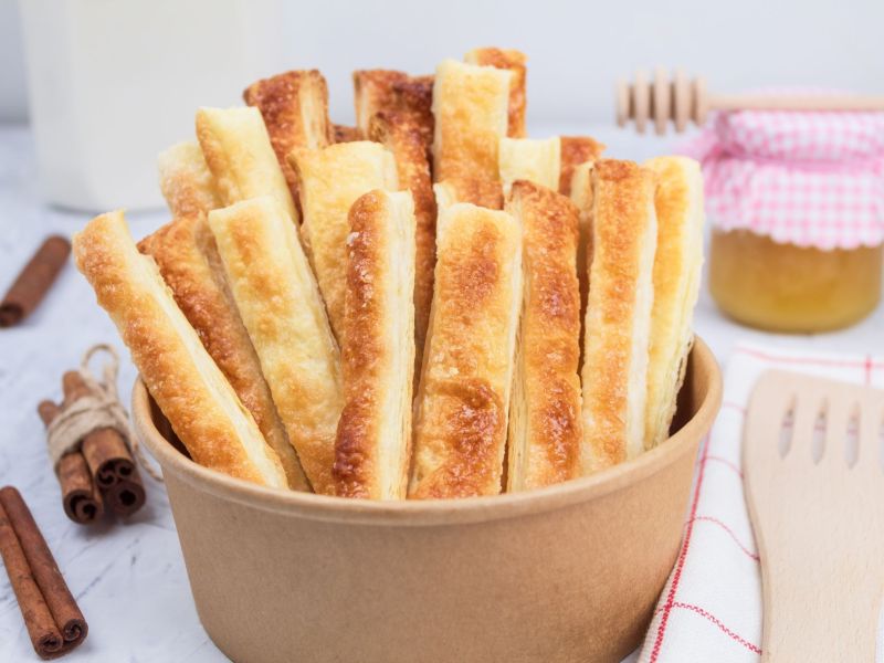 Puff pastry pudding sticks, served in a bowl. Cinnamon sticks and a wooden scraper lie next to it as decoration. A jam jar can be seen in the background.