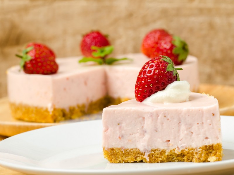 A piece of strawberry mousse cake, garnished with a strawberry, on a plate, with the cut cake in the background.