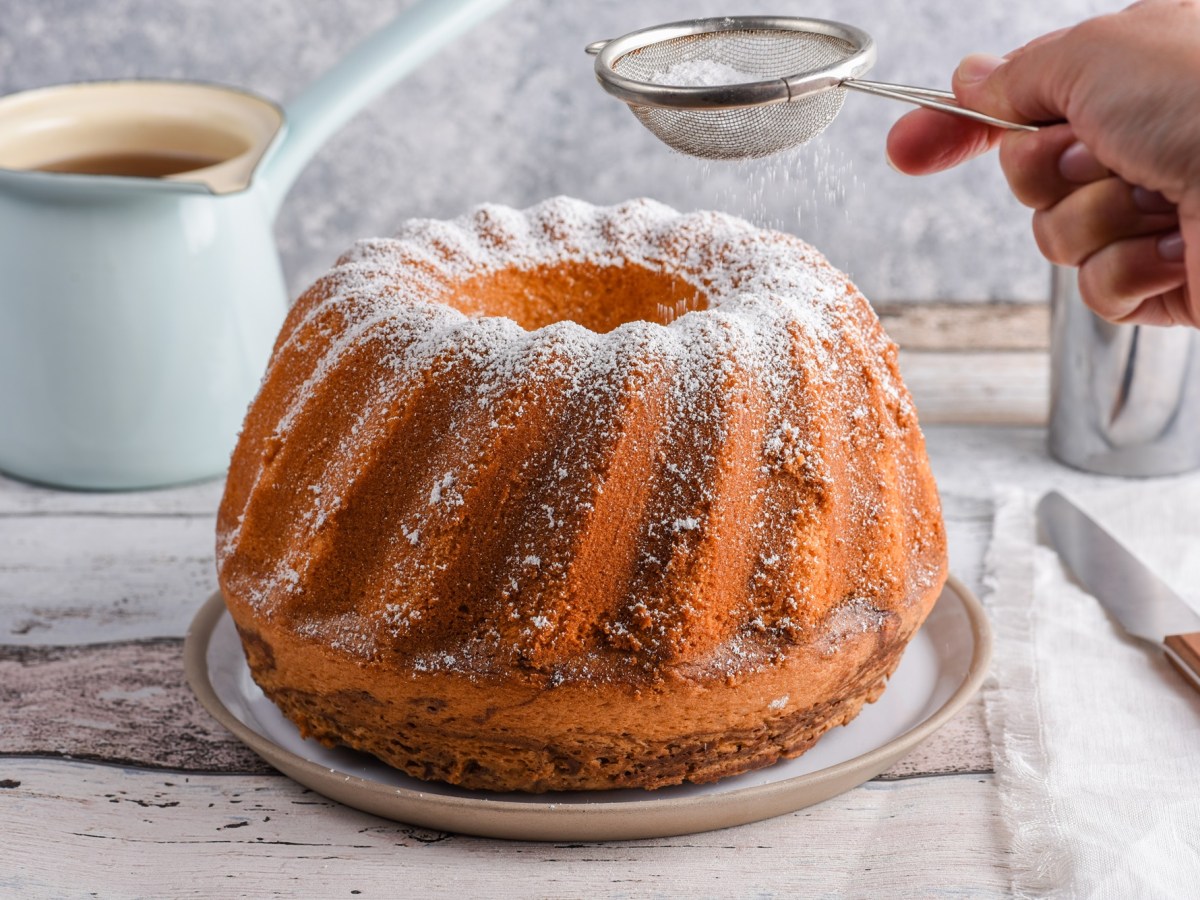 An amaretto bundt cake on a plate being dusted with powdered sugar. A knife rests on a piece of cloth next to the cake. A filled glass stands in the background.