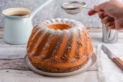 An amaretto bundt cake on a plate being dusted with powdered sugar. A knife rests on a piece of cloth next to the cake. A filled glass stands in the background.