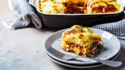 A piece of Greek Gyro Potato Casserole lies in front of a casserole dish with more casserole in it.