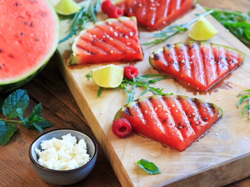 Grilled watermelon slices on a wooden board. Next to it is a bowl of feta.