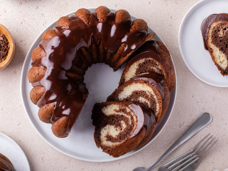Top view of a sliced Nutella marble cake in a Bundt pan. The cake is on a white plate. Next to it on the right is another plate with a piece of marble cake with Nutella. To the left of it is a bowl of chocolate chips and there are some cake forks on the work surface.