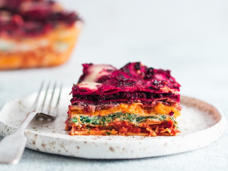A piece of vegan rainbow lasagna on a plate. The rest can be seen in the background.
