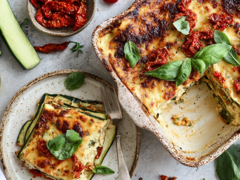 Overhead view of a baking dish with zucchini-feta lasagna. A piece of it lies on a plate next to it. Fresh ingredients such as zucchini, a bowl of sun-dried tomatoes, a glass jar with olive oil, fresh basil are scattered around.
